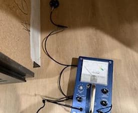 Blue electronic device on a wooden floor with wires, likely for measuring or diagnostics.