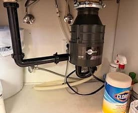 Under-sink garbage disposal unit with plumbing connections and cleaning supplies nearby.