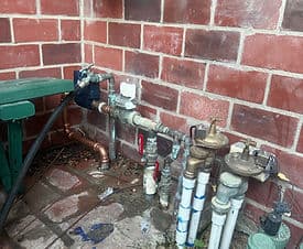 Pipe system with valves and water meter against a red brick wall, showing plumbing details.