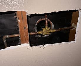 Plumbing installation behind wall with copper pipes and valve visible in cutout opening.